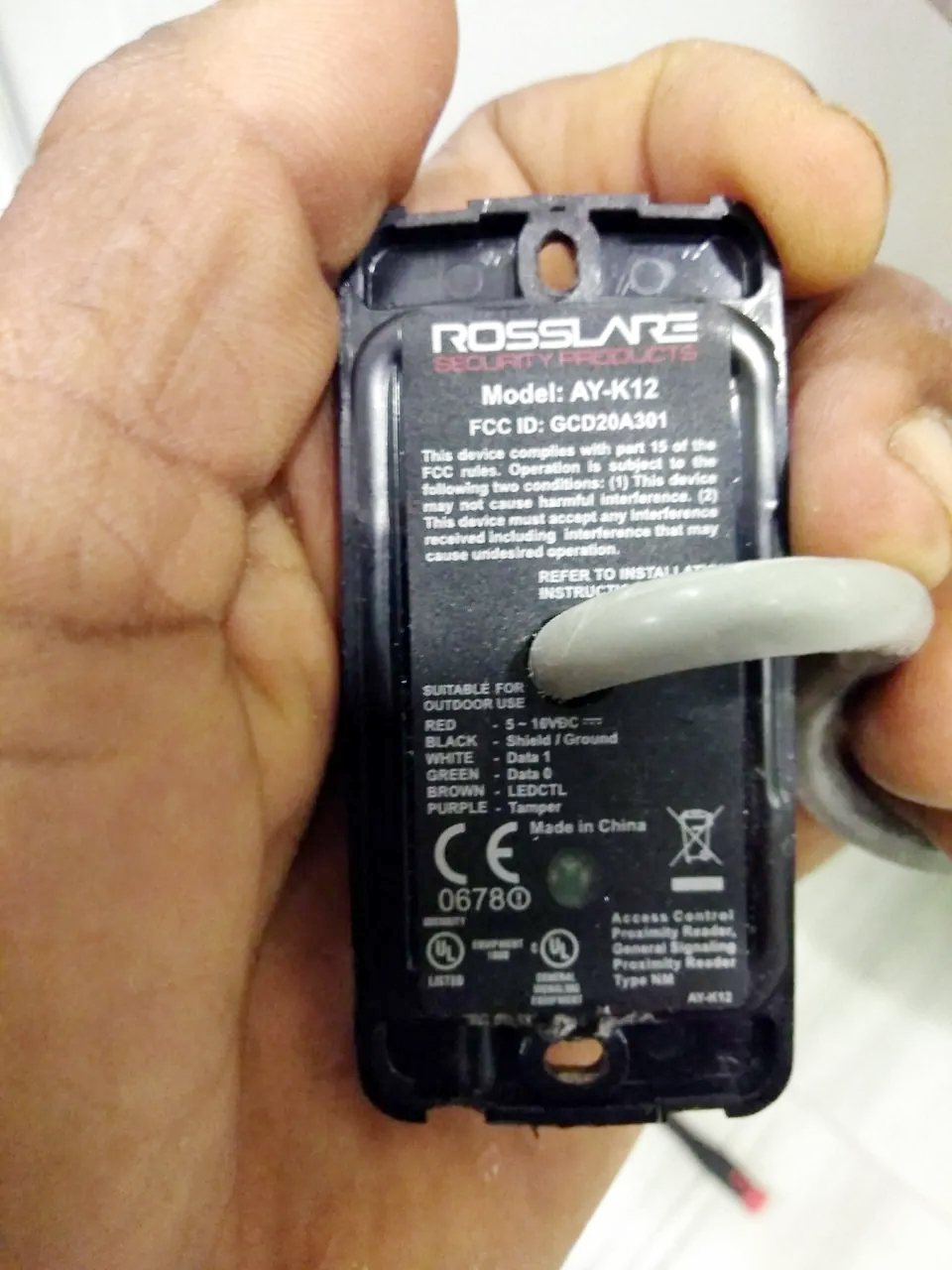 A hand holding a Rosslare access reader, showing its cable and equipment label.